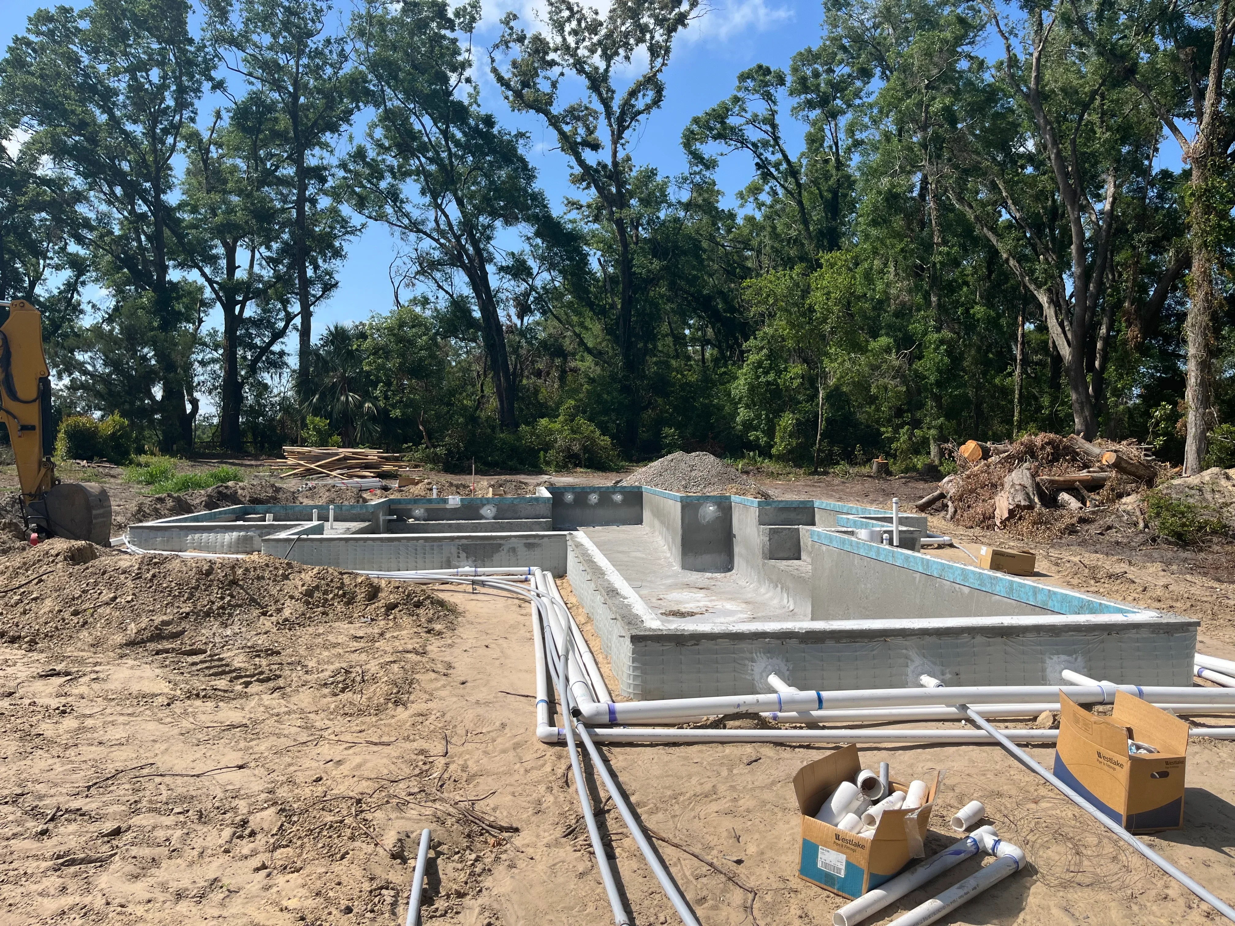 Concrete pool foundation under construction with formed walls, plumbing lines, and surrounding excavation