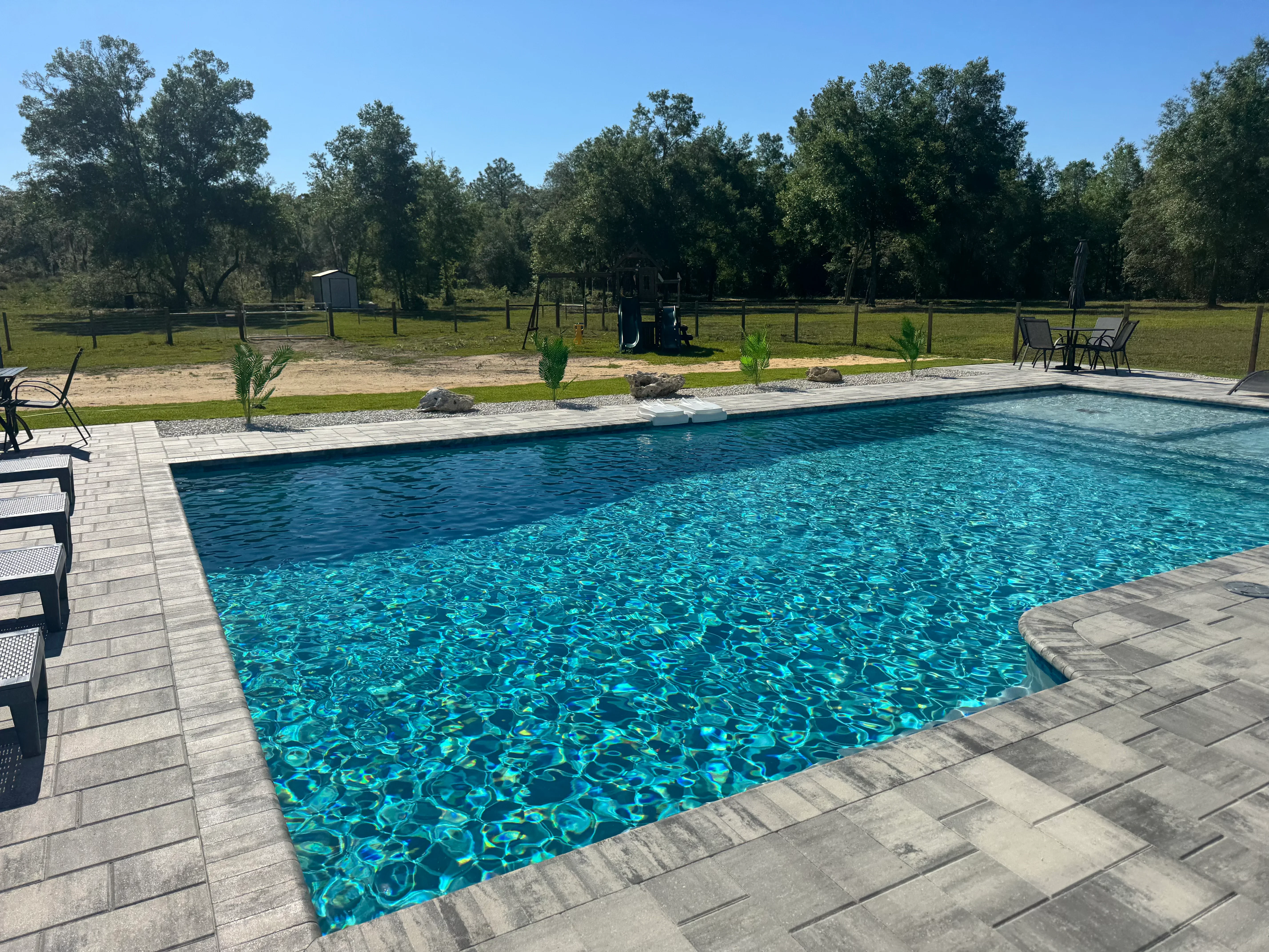 Rectangular swimming pool with gray paver deck, lounge chairs, and natural landscape backdrop
