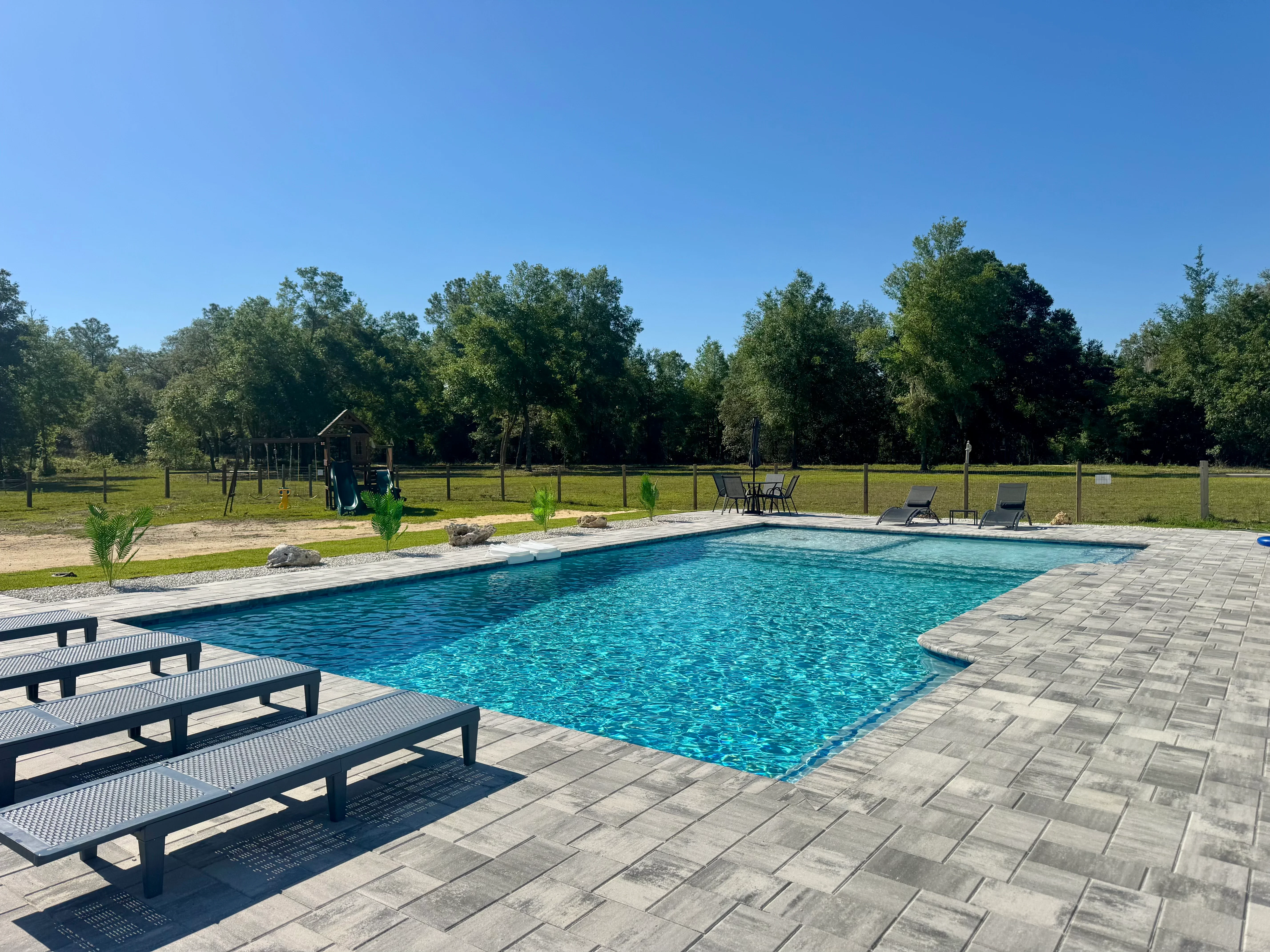 Rectangular swimming pool with gray paver deck, lounge chairs, and manicured landscaping