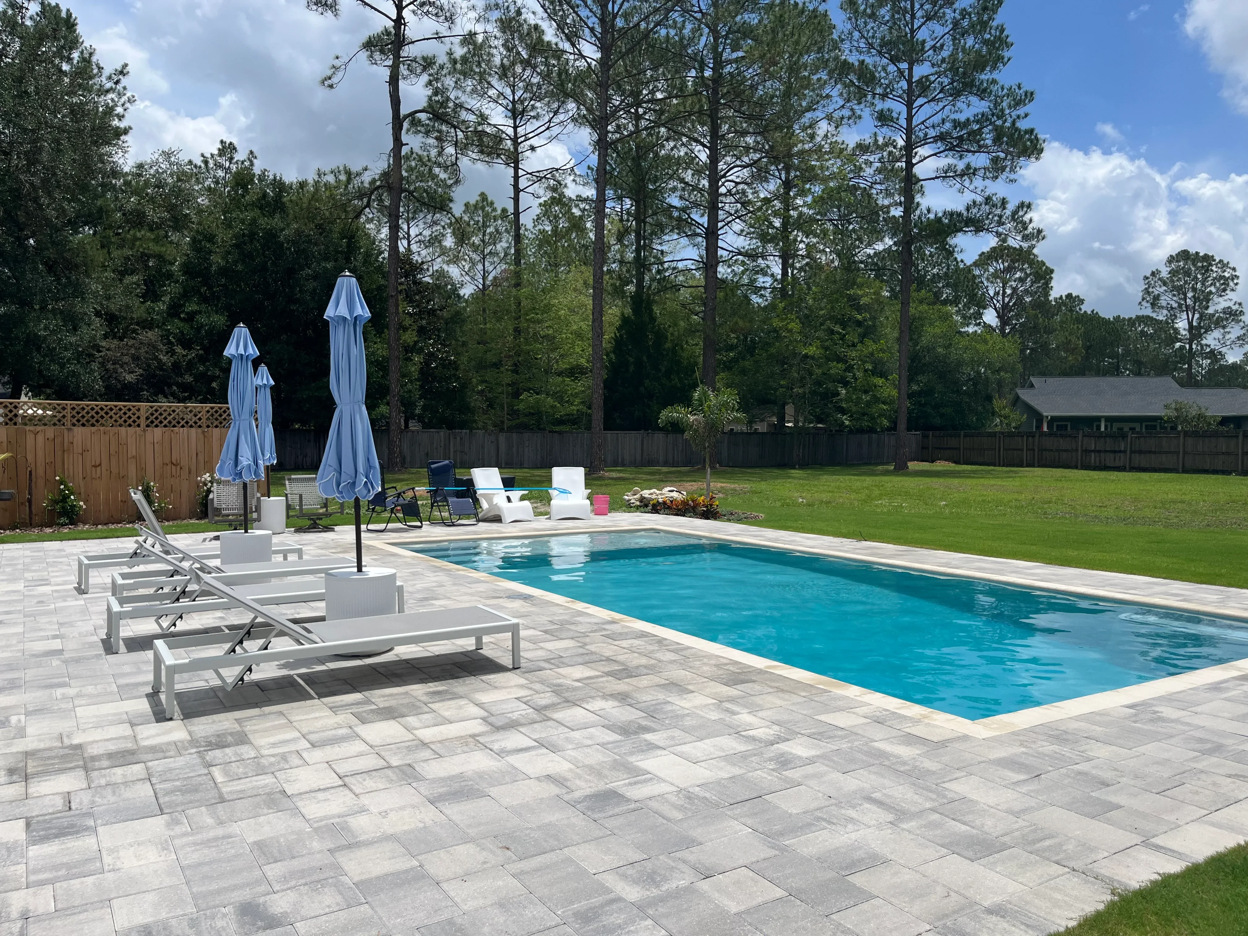 In-ground pool with gray paver deck, lounge chairs, and blue umbrellas in wooded backyard