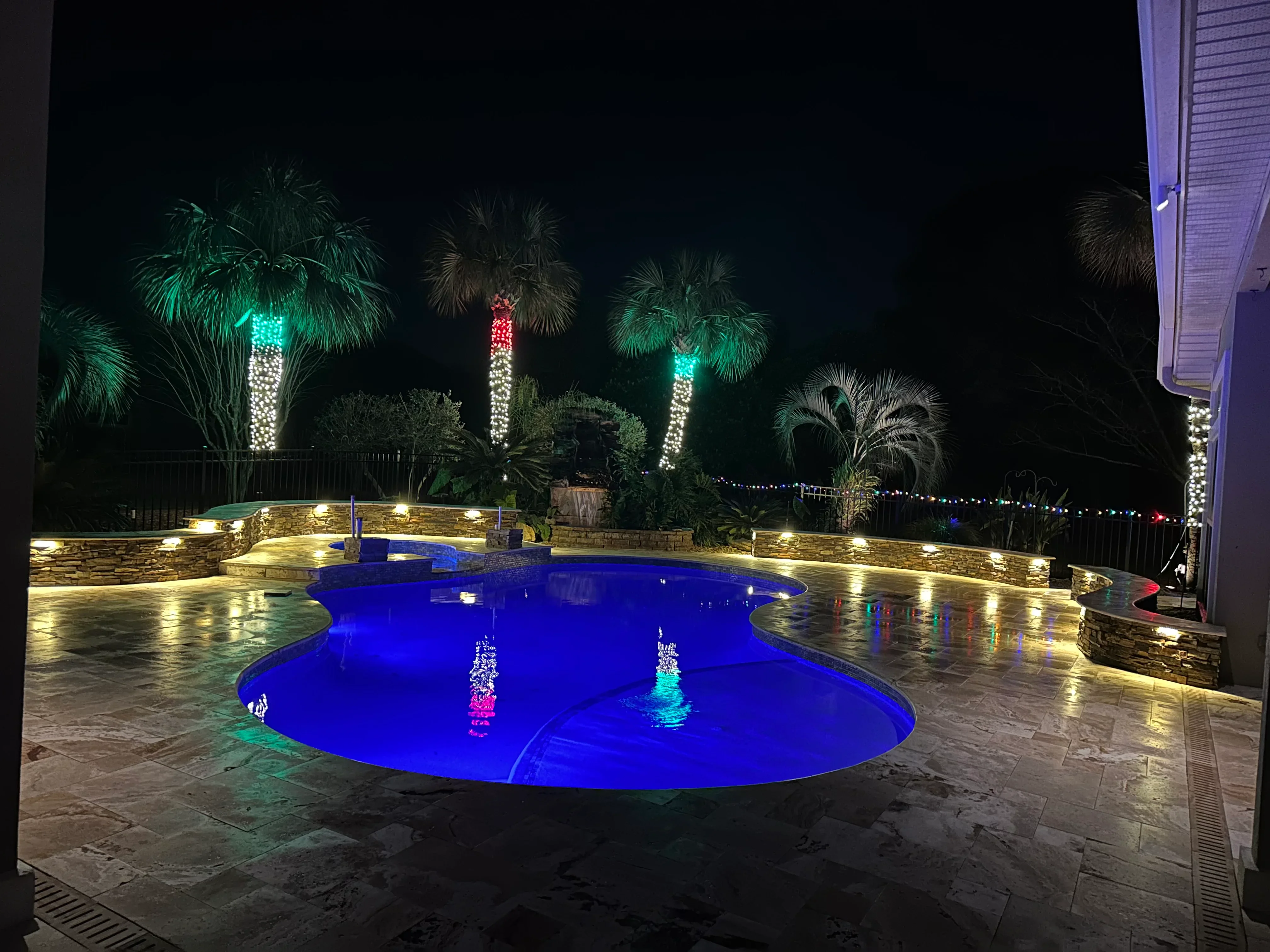 Illuminated resort-style pool with blue LED lighting, tropical palms, and stone patio at night