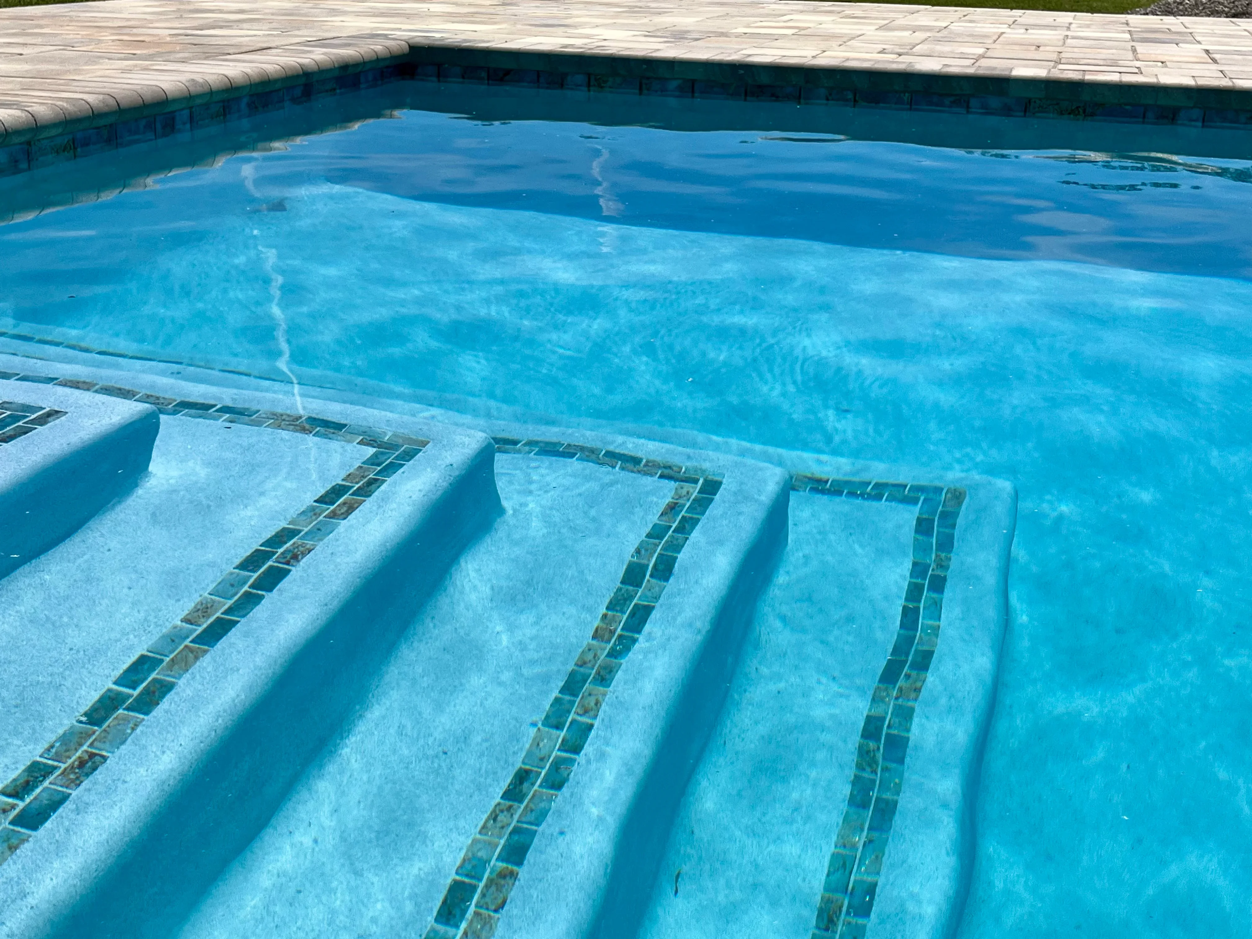 Bright blue residential pool with three concrete steps featuring dark tile accents and surrounding paver deck